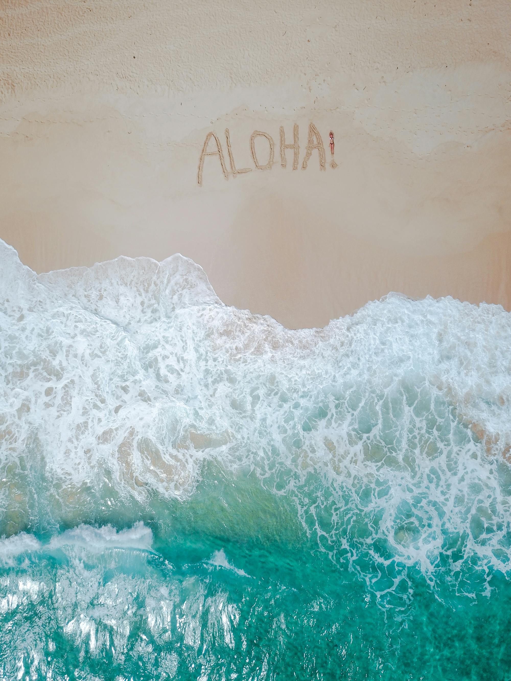 A serene aerial shot of a tropical beach with 'Aloha' written in the sand and waves crashing ashore.